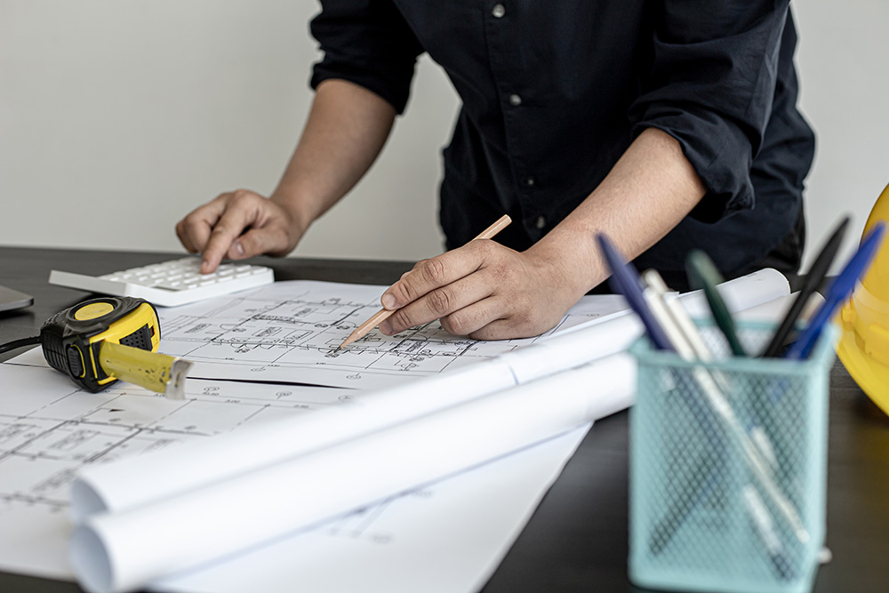 Person using a pencil to mark architectural blueprints and a cabinet measurement guide on a desk.