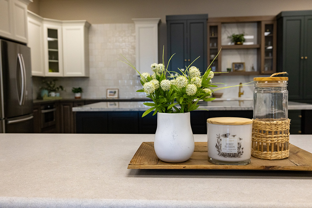 A white vase with flowers, a Honey Blossom candle, and a jar on a wooden kitchen cabinet tray.