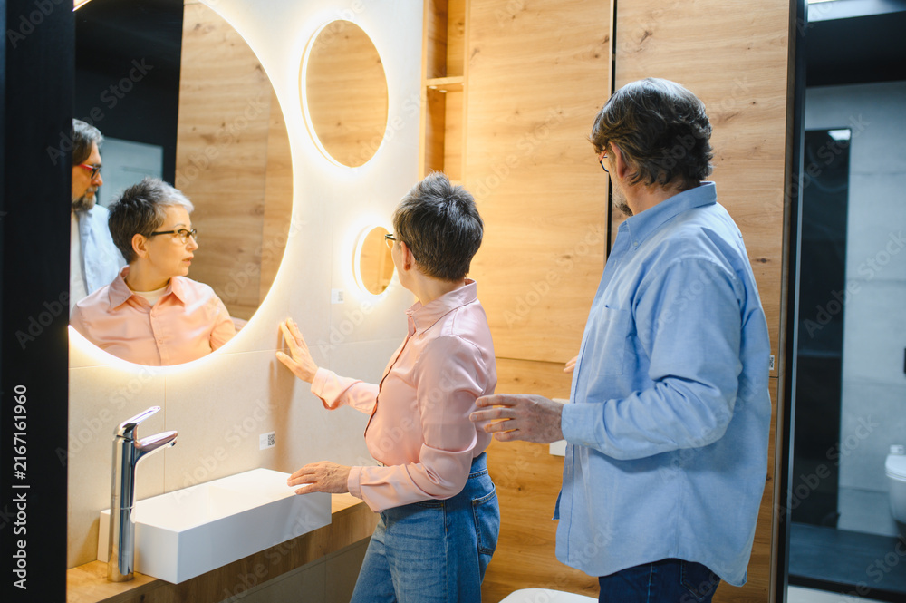 A man and woman examine a lighted circular bathroom mirror near bathroom cabinets in a showroom.