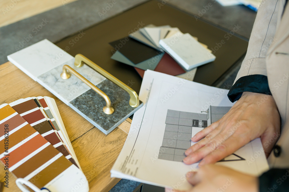 Hands hold a floor plan on a desk with material samples and gold kitchen cabinet pulls.