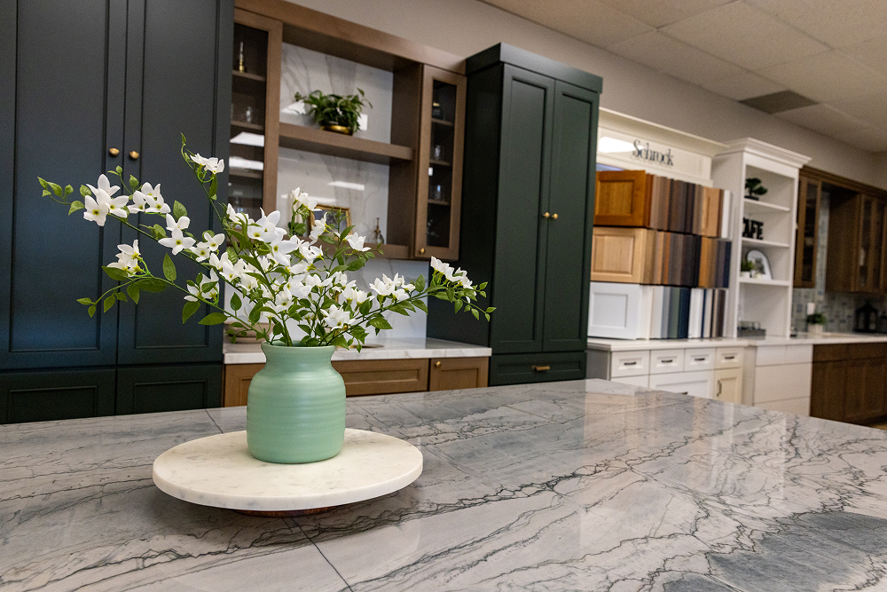 White flowers in a green vase on a grey marble kitchen island with dark green background cabinets at the Bay Area Cabinet Supply showroom in the Bay Area.